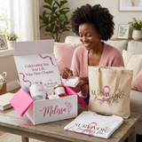 A smiling woman sits on a couch in a living room, opening a white gift box and holding a small item while surrounded by breast cancer awareness merchandise, including a tote bag, a folded T-shirt, and a branded box.