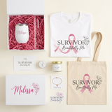 A curated gift set featuring a white t-shirt and canvas tote bag with a pink ribbon graphic and the text 'SURVIVOR Beautifully Me,' a white insulated tumbler in pink shredded paper inside an open box labeled 'Melissa,' a small candle, jewelry card, and matching branded cards and boxes on a light cream background.