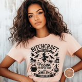 A woman with long, wavy brown hair wearing a light pink graphic t-shirt. The t-shirt features a black printed design with the words 'Bitchcraft - The Art of Pissing People Off and Smiling Sweetly' along with witch illustrations like a broomstick, potion bottle, and house.