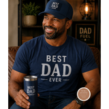 Man wearing a navy 'Best Dad Ever' shirt and cap, holding a matching mug, in a cozy indoor setting.