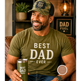 Man wearing a 'Best Dad Ever' shirt and cap, holding a 'Best Dad Ever' mug in a cozy room.