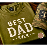 Green t-shirt with 'Best Dad Ever' text and camouflage cap on a wooden surface