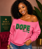 A smiling woman with curly black hair models a bright pink off-the-shoulder sweatshirt featuring the word 'DOPE' in green puff vinyl 'SINCE 1908' printed below. She is wearing blue jeans and gold hoop earrings, standing in a styled room with a pink wall, a green sign reading 'MOKA QUEENZ', and a potted plant.