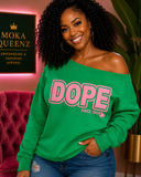 A woman wearing a kelly green off-the-shoulder sweatshirt with 'DOPE SINCE 1998' printed in pink across the front, paired with blue jeans in a styled indoor setting