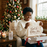 Woman opening her God Is Within Her Christmas Gift Box by the tree with the matching shirt, cup, and tote