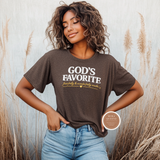 A woman wearing a Heather dark brown graphic t-shirt with the text 'GOD'S FAVORITE' in white and 'feel fully & wonderfully made' in gold script, paired with light blue jeans, standing in front of a rustic wall with dried grass.