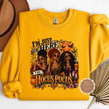 A folded yellow sweatshirt with a colorful graphic print of three witches and the text 'I'M JUST HERE FOR THE HOCUS POCUS', styled on a white background with a brown hat, dried palm leaves, and a circular 'Moka Queenz' badge.