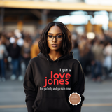 A woman wearing a black hoodie with the text 'I got a love jones Fo' ya body and ya skin tone' in red and white, standing in a blurred outdoor crowd setting. A small circular 'Abuja Cap Hits' logo is visible on the lower right of the hoodie.