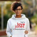 A woman wearing a white hoodie with the text 'I got a love Jones' and 'Fo' ya body and ya skin tone' printed on the front, standing outdoors against a blurred green background.