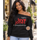 A woman wearing a black off-the-shoulder sweatshirt with the text 'I got a love Jones' in red and white, and 'Fo' ya body and ya skin tone' below it, standing outdoors in front of a storefront.