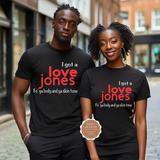 A man and a woman standing together on a city street, both wearing matching black graphic T-shirts with red and white text that reads 'I got a love jones' and a smaller line below. The woman's shirt has a visible 'More Queenz' badge overlaid near the lower front.
