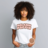 A woman with curly dark hair wearing a white graphic t-shirt that reads 'MELANIN POPPIN' in a pink and brown bubble font, paired with light-wash jeans, against a solid gray studio background.