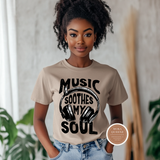Woman wearing a beige t-shirt with 'Music Soothes My Soul' design, standing indoors with plants in the background.