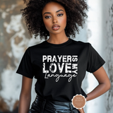 A woman with curly dark hair wearing a black short-sleeve graphic T-shirt with the phrase 'PRAYERS LOVE MY Language' in distressed white lettering. She is posing against a softly blurred, light-toned indoor background, with a small circular 'Moka Queenz' brand badge overlaid near the bottom right.