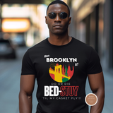 A man wearing a black graphic t-shirt with a colorful print of the Brooklyn Bridge and the text 'Where BROOKLYN AM? DO OR DIE BED-STUY TIL MY CASKET FLY!!!' in an outdoor blurred street setting.