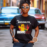 A woman wearing a black graphic t-shirt with 'BROOKLYN AT? BROWNSVILLE NEVER DAN - NEVER WILL' and a city skyline graphic, standing in front of a blurred street background with vintage cars.