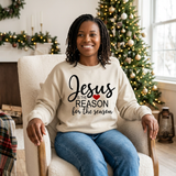 Black woman seated in a beige Jesus Is the Reason for the Season sweatshirt with black text and a red heart.