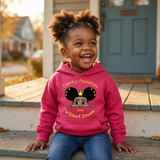 Laughing Black toddler girl with afro puffs on a front porch wearing a raspberry pink I Am My Ancestors Wildest Dream hoodie with the name Ava