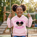 Laughing Black girl on a playground swing wearing a light pink I Am My Ancestors Wildest Dream hoodie with the name Ava