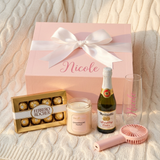 Spa Day gift box tied with a white satin bow beside the Ferrero Rocher 8-pack, Champagne Toast candle, mini Martinelli's, Nicole flute, and pink fan on a knit blanket
