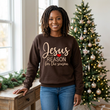 Black woman standing by a window in a brown Jesus Is the Reason for the Season sweatshirt with tan text.