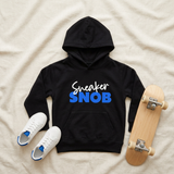 Black Sneaker Snob kids hoodie flat lay with blue print, white sneakers, and a skateboard