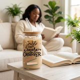 Just One More Chapter iced coffee glass with bamboo lid and glass straw beside an open book in a cozy reading nook.