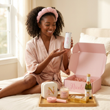 Smiling Black woman in a blush robe and pink spa headband holding her Nicole champagne tumbler beside her open Spa Day gift box