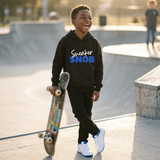 Laughing Black boy with a skateboard at a skate park wearing a black Sneaker Snob hoodie with blue print