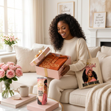 Smiling woman on a cream sofa opening her Special Girl Gift Box with the hoodie inside, the affirmation tumbler on the coffee table, and the Special Girl tote on her shoulder