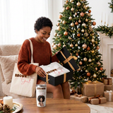 Woman opening her Brown Skin Girl Christmas Gift Box by the tree with the sweatshirt, white matching tumbler, and tote