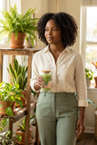 Woman holding a boho botanical iced matcha glass beside a plant shelf in a sunlit home.