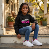 Smiling Black girl with braids on a front porch wearing a black Sneaker Snob hoodie with pink print and white sneakers