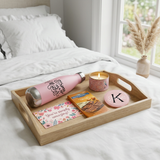 Strong As A Mother self-care tray with the water bottle, lit candle, Ghirardelli bar, monogram mirror, and postcard on white bedding