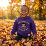 Smiling Black toddler boy sitting in golden fall leaves wearing a purple I Am My Ancestors Wildest Dream hoodie with the name Dante