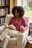 Woman reading in a cozy armchair while holding a Hot Girls Read glass filled with iced matcha.
