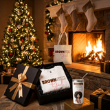 Brown Skin Girl Christmas Gift Box by a fireplace and Christmas tree with the sweatshirt, white matching tumbler, and tote