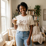 Smiling Black woman wearing the God Is Within Her shirt with jeans, holding the matching glass can cup