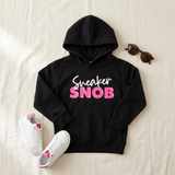 Black Sneaker Snob kids hoodie flat lay with pink print, white sneakers, and sunglasses