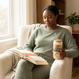 Woman reading in a sunlit armchair while holding a Just One More Chapter iced coffee glass with bamboo lid and glass straw.