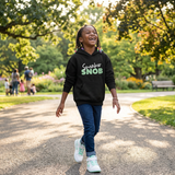 Laughing Black girl walking in a sunny park wearing a black Sneaker Snob hoodie with mint print and white sneakers