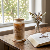 God Is Within Her frosted glass can filled with iced coffee beside an open Bible by a window
